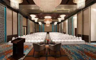 Long presentation and banquet hall with ornate chandeliers, a podium and sitting area for two panelists at the front, at the Hilton Jaipur, India.