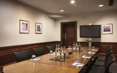 A large glossy wooden table with glass water carafes and seating for 8 participants with large television mounted on wall.