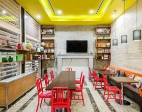 A long rectangular wooden table in the middle with red chairs. Several other smaller tables for 4 to work, eat, socialize at. Yellow lit ceiling and white fireplace mantle frames the room.