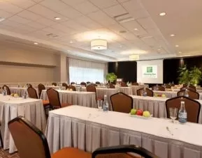 Meeting room with numerous tables and seating arranged to face a lectern, beside which is a projector screen.
