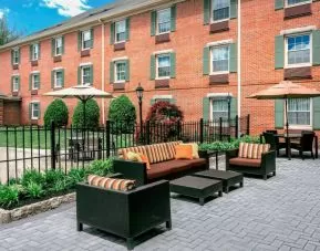 Sonesta Select Tinton Falls Eatontown’s patio is furnished with shaded tables and chairs, in addition to sofa and armchair seating, with greenery nearby.
