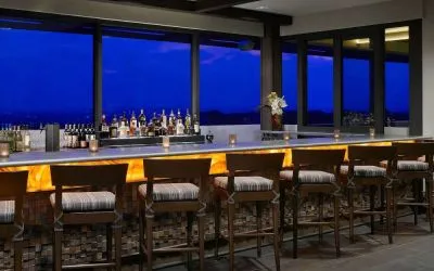 The hotel bar has traditional tall bar stools, a hard floor, a wide array of beverages, and large windows.