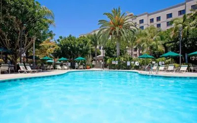 The outdoor pool at Sonesta ES Suites Anaheim Resort Area features nearby sun loungers and shaded tables and chairs, with numerous trees close by.