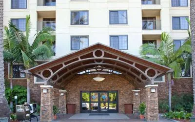 The hotel's exterior features a covered entrance, with potted plants and large trees and other greenery to either side.