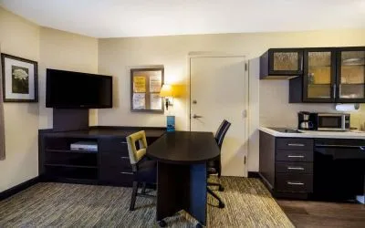 Sonesta Simply Suites St Louis Earth City guest room workspace, furnished with desk and chairs, with TV and kitchen nearby.