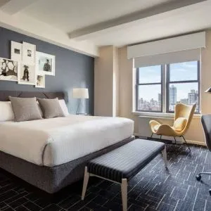 Double bed guest room in The Shelburne Sonesta New York, featuring a window, art on the wall, and a workspace desk, chair, and lamp.