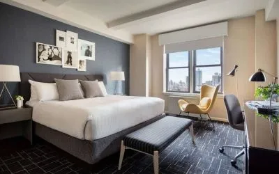 Double bed guest room in The Shelburne Sonesta New York, featuring a window, art on the wall, and a workspace desk, chair, and lamp.