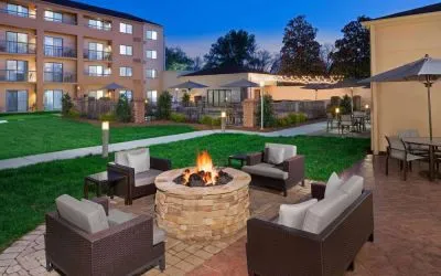 Sonesta Select Raleigh Durham Airport Morrisville’s fire pit has armchair and sofa seating, plus coffee tables, next to it, with shaded tables and chairs nearby.