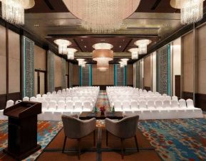 Long presentation and banquet hall with ornate chandeliers, a podium and sitting area for two panelists at the front, at the Hilton Jaipur, India.
