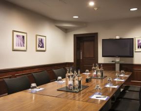 A large glossy wooden table with glass water carafes and seating for 8 participants with large television mounted on wall.