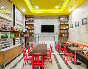 A long rectangular wooden table in the middle with red chairs. Several other smaller tables for 4 to work, eat, socialize at. Yellow lit ceiling and white fireplace mantle frames the room.