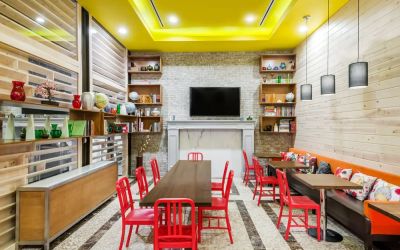 A long rectangular wooden table in the middle with red chairs. Several other smaller tables for 4 to work, eat, socialize at. Yellow lit ceiling and white fireplace mantle frames the room.