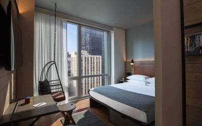Moxy NYC Downtown double bed guest room, with large television, floor-to-ceiling window, and desk for working.