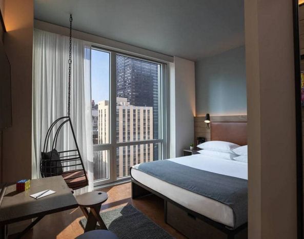 Moxy NYC Downtown double bed guest room, with large television, floor-to-ceiling window, and desk for working.