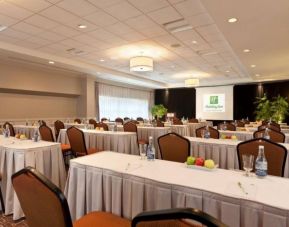 Meeting room with numerous tables and seating arranged to face a lectern, beside which is a projector screen.