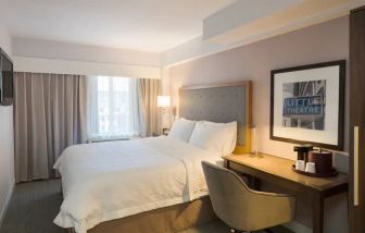 Cozy hotel room with beige curtains, wall-mounted television, white linen over full-size bed, with adjacent desk complete with coffeemaker and comfortable leather chair.