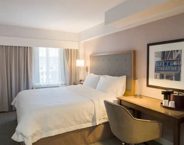 Cozy hotel room with beige curtains, wall-mounted television, white linen over full-size bed, with adjacent desk complete with coffeemaker and comfortable leather chair.