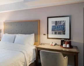 Full-size bed with olvie bed header, adjacent hotel room workspace with seude chair, coffeemaker, phone and desk lamp.