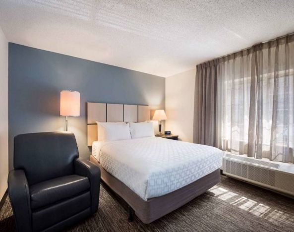 Double bed guest room in Sonesta Simply Suites Nashville Brentwood, featuring armchair, bedside lamp, and window.