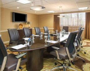 Hotel meeting room, furnished with long table, 10 swivel chairs, chocolate hues, and a wall-mounted television.