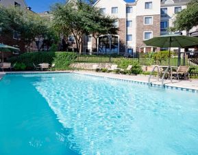 Sonesta ES Suites Austin The Domain Area’s outdoor pool features tables and chairs by the side, some of which are shaded, and nearby trees.