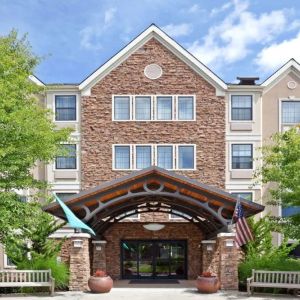 The hotel’s exterior has a covered entrance, wooden benches, and pleasant greenery including flowers and trees.