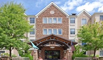 The hotel’s exterior has a covered entrance, wooden benches, and pleasant greenery including flowers and trees.