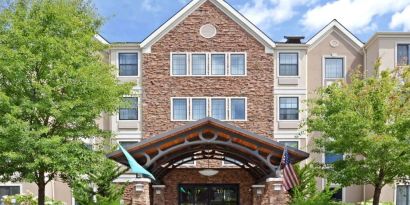The hotel’s exterior has a covered entrance, wooden benches, and pleasant greenery including flowers and trees.