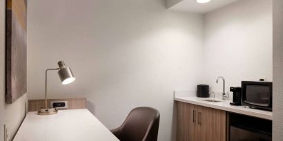 Guest room workspace in Sonesta Select Seattle Renton, including desk, chair, and lamp, with kitchenette nearby.