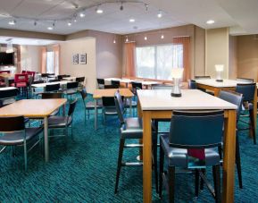 The hotel’s breakfast area is furnished with a carpet, mix of table sizes, large windows, and a TV.