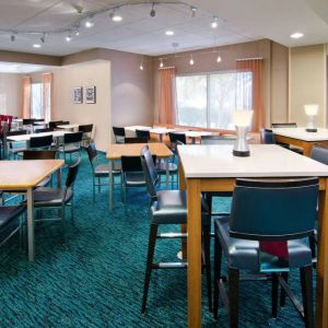 The hotel’s breakfast area is furnished with a carpet, mix of table sizes, large windows, and a TV.