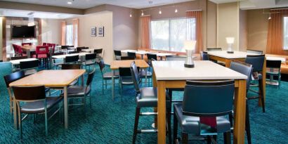 The hotel’s breakfast area is furnished with a carpet, mix of table sizes, large windows, and a TV.