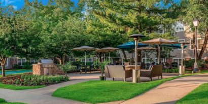 Sonesta ES Suites Parsippany Morris Plains’ barbecue area is furnished with tables and chairs, plus armchair seating, amid numerous trees.