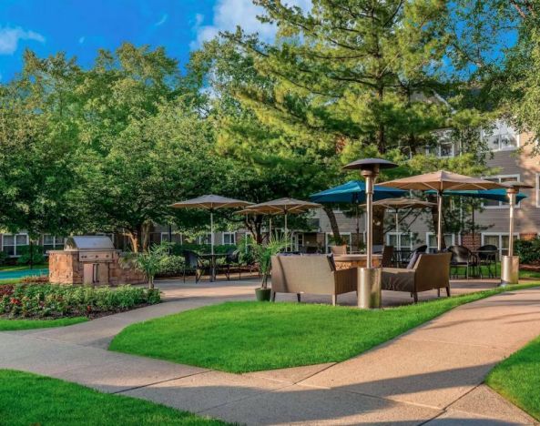 Sonesta ES Suites Parsippany Morris Plains’ barbecue area is furnished with tables and chairs, plus armchair seating, amid numerous trees.