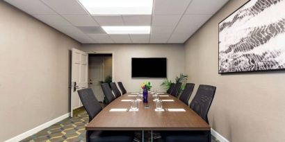 Hotel meeting room, featuring long wooden table, eight swivel chairs, and a large, widescreen TV on the wall.