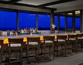 The hotel bar has traditional tall bar stools, a hard floor, a wide array of beverages, and large windows.