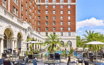 The hotel patio features tables and chairs, some shaded, overlooking the outdoor pool.