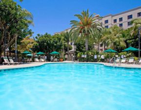 The outdoor pool at Sonesta ES Suites Anaheim Resort Area features nearby sun loungers and shaded tables and chairs, with numerous trees close by.