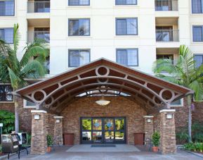 The hotel's exterior features a covered entrance, with potted plants and large trees and other greenery to either side.