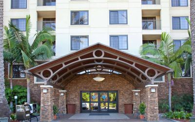 The hotel's exterior features a covered entrance, with potted plants and large trees and other greenery to either side.