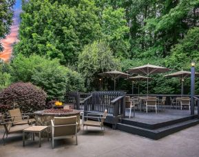 Sonesta Charlotte Executive Park’s patio includes shaded tables and chairs, and a fire pit surrounded by chairs and coffee tables, with abundant greenery nearby.