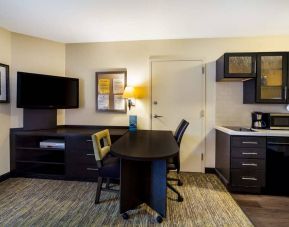 Sonesta Simply Suites St Louis Earth City guest room workspace, furnished with desk and chairs, with TV and kitchen nearby.