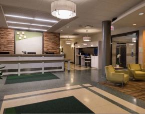 The hotel lobby has yellow armchairs, potted plants, and a business center workstation, in addition to the reception desk.