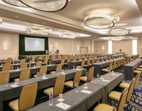 Hotel meeting room, furnished with tables and seating for dozens of attendees, arranged in a classroom style facing a large projector screen.