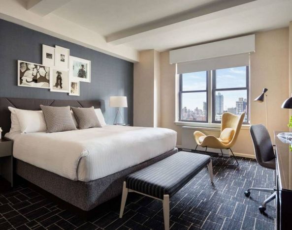 Double bed guest room in The Shelburne Sonesta New York, featuring a window, art on the wall, and a workspace desk, chair, and lamp.