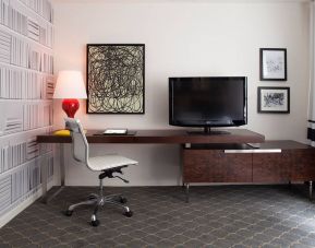 Guest room workspace in The Fifty Sonesta Select New York, featuring desk, chair, and lamp, with art on the wall and a nearby TV.