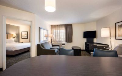 Sonesta Simply Suites Detroit Troy double bed guest room, including living area with table and chairs, sofa, TV, and window.