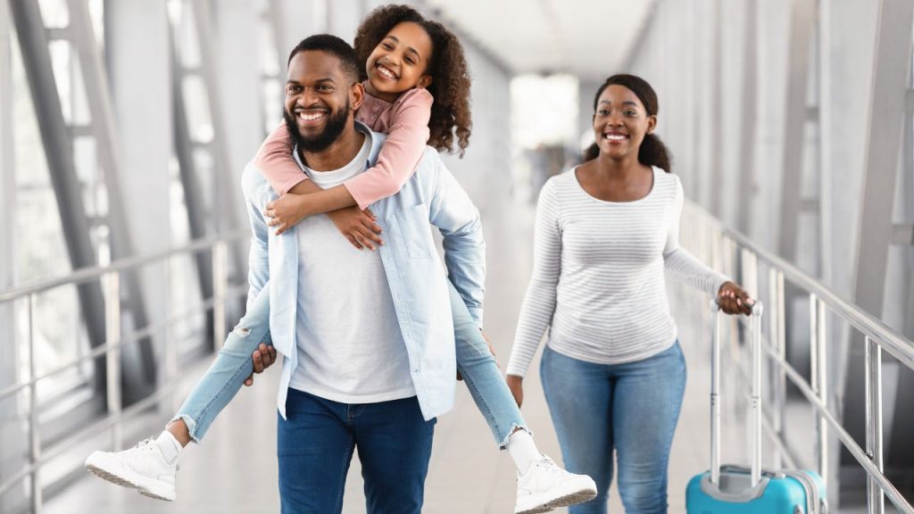 happy parents at airport: lady at airport rolling luggage while man carries daughter on piggy back