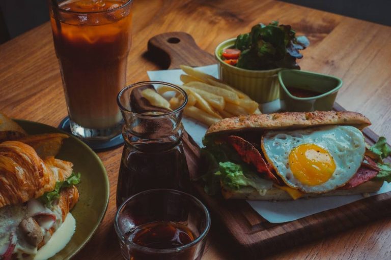 Wooden table with sunny side egg and ham sandwich, french fries, iced coffee and soda.