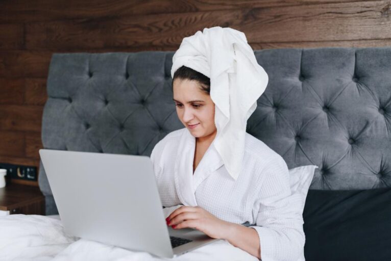 Woman works on laptop from hotel bed after shower.
